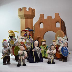 Könnte beinhalten: Eine Sammlung handgefertigter Puppen im mittelalterlichen Stil, darunter Ritter, ein Narr und ein König, vor einer Holzburg. Die Puppen tragen detaillierte Kostüme mit Schilden, Speeren und Kronen. Die Burg ist aus hellbraunem Holz.