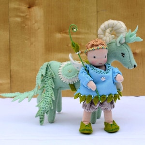 Puede incluir: Una muñeca hecha a mano con una túnica azul, adornos en forma de hojas y pantalones cortos marrones está junto a un caballo de fieltro. La muñeca tiene una corona y sostiene un bastón verde. El caballo es verde claro con una silla blanca.