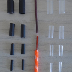 May include: Assortment of black and clear plastic cord ends in various sizes. The cord ends are arranged in rows on a light wood surface. A cord with a brown and orange color scheme is in the center of the image.