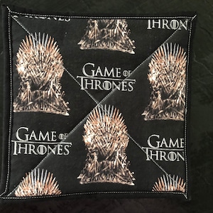May include: Square black fabric coaster featuring the Iron Throne and the words "Game of Thrones" in white. The coaster has white stitching around the edges.