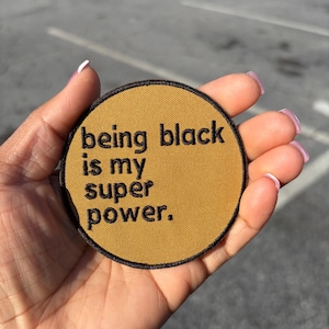 May include: A round, mustard-colored patch with black embroidered text that reads "being black is my super power." The patch has a black border and is held in a hand against a blurred background.