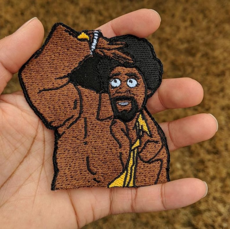 May include: Embroidered patch featuring a cartoon man with an afro, wearing a yellow collared shirt. The patch is brown, black, and yellow, with a detailed design. The man is looking up with a surprised expression. The patch is held in a hand.