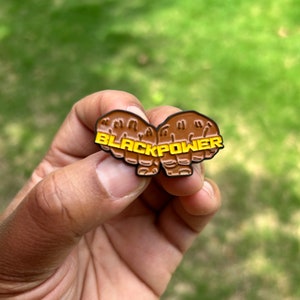 Pode incluir: Um alfinete de esmalte marrom e amarelo em forma de dois punhos. O alfinete tem as palavras "BLACK POWER" em letras amarelas. O alfinete é segurado em uma mão contra um fundo verde desfocado.