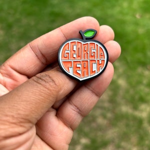 May include: A peach-shaped charm with the words "GEORGIA PEACH" in orange and white, outlined in black. The charm has a green leaf and is held against a blurred green background.