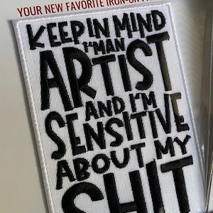 Op de afbeelding: Wit rechthoekig opstrijkpatch met zwart geborduurde tekst. De tekst luidt: "KEEP IN MIND I'M AN ARTIST AND I'M SENSITIVE ABOUT MY SHIT." De patch bevat ook de tekst "A HOUSE CALLED HUE" en "YOUR NEW FAVORITE IRON-ON PATCH".