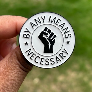 Pode incluir: Um alfinete redondo de esmalte branco com uma borda preta. O alfinete apresenta um punho negro no centro, rodeado pelas palavras "BY ANY MEANS NECESSARY" em texto preto. Duas estrelas pretas ladeiam o texto.