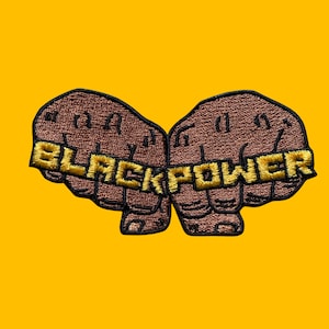 Pode incluir: Patch bordado com dois punhos cerrados castanhos com a inscrição dourada "BLACK POWER". O patch está sobre um fundo amarelo brilhante.