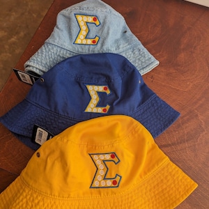 Puede incluir: Tres sombreros de pescador en mezclilla, azul marino y amarillo. Cada sombrero presenta una letra griega Sigma amarilla bordada con puntos blancos y detalles rojos. Los sombreros se exhiben sobre una superficie de madera.