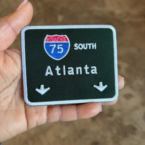 May include: A dark green patch with white border featuring the text "I-75 SOUTH Atlanta" and a logo of a blue and red Interstate 75 shield. Two white arrows point downwards. The patch is held in a hand.