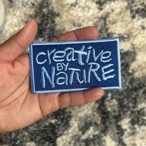May include: A rectangular blue patch with the embroidered text "creative BY nature" in white. The patch has a light blue border and is likely designed to be sewn onto clothing or accessories. The patch is held in a hand.