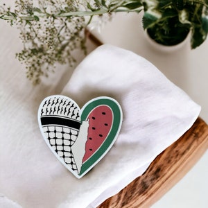 May include: A heart-shaped sticker with a black and white keffiyeh pattern on one side and a watermelon slice on the other. The watermelon slice is green and red with black seeds.