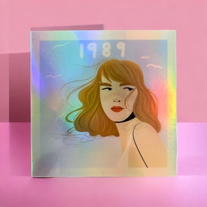 May include: Square holographic sticker featuring an illustration of a woman with flowing auburn hair and red lipstick. The word "1989" is printed above the woman's head in a light blue font. The background has a light blue and white gradient.