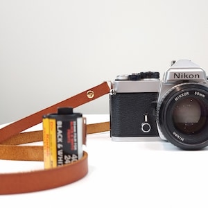 May include: A silver Nikon film camera with a black Nikkor 50mm f/1.4 lens. The camera is attached to a brown leather strap and a roll of black and white film is next to the strap.
