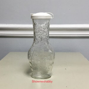 May include: Clear glass pitcher with a textured surface and a white plastic lid. The pitcher has a curved shape and a narrow neck. The text "Showmeshabby" is visible at the bottom of the image.