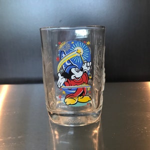 Puede incluir: Un vaso de vidrio transparente con un diseño de Mickey Mouse. Mickey lleva una túnica roja y azul y un sombrero de mago. Está sosteniendo una varita mágica y estrellas lo rodean. El vaso tiene el texto "Disney" y "2000".