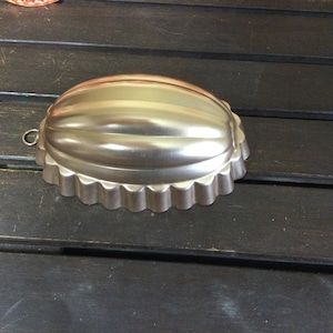 Op de afbeelding: Een zilverkleurige, gegroefde metalen vorm met een afgeronde, geribbelde bovenkant en een geschulpte rand. Het heeft een kleine lus om op te hangen. De vorm is waarschijnlijk bedoeld voor het maken van gelatinedesserts of andere gevormde voedingsmiddelen.