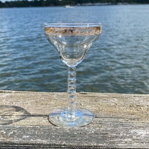Puede incluir: Una copa de cóctel transparente con borde dorado y tallo decorativo. La copa se encuentra sobre un fondo de agua y una superficie de madera. La copa está diseñada para servir cócteles. El tallo tiene una serie de hendiduras circulares.