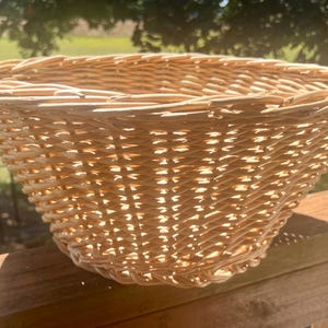 May include: A light brown, woven wicker basket with a flared shape. The basket is made of tightly woven, natural-colored wicker and sits on a wooden surface. The basket's open design and sturdy construction make it suitable for various uses.