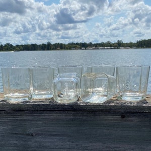 May include: Six clear glass tumblers, some with a square shape, are lined up on a wooden surface. The glasses are empty and appear to be clean.