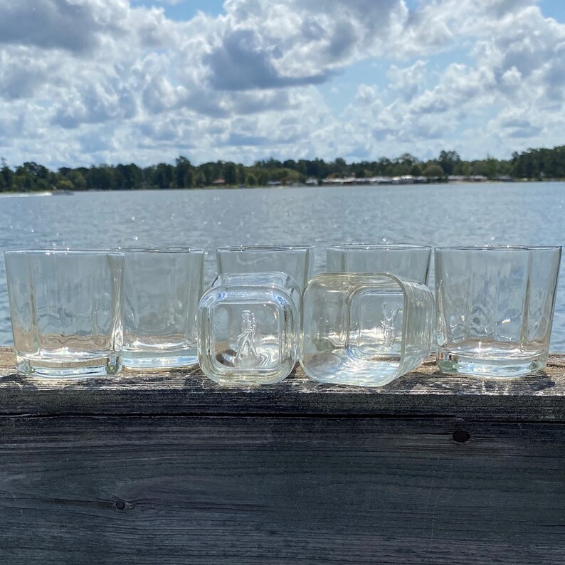 May include: Six clear glass tumblers, some with a square shape, are lined up on a wooden surface. The glasses are empty and appear to be clean.