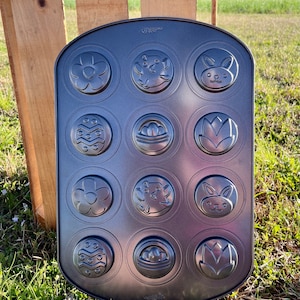 Könnte beinhalten: Eine dunkelgraue Backform mit 12 kreisförmigen Formen. Jede Form zeigt ein anderes Ostermotiv, darunter Blumen, Hasen, Eier und Tulpen. Die Form ist aus Metall und zum Backen bestimmt. Der Markenname "Wilton" ist sichtbar.