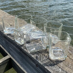 May include: Six clear glass tumblers with a textured surface, arranged in a row on a wooden surface.