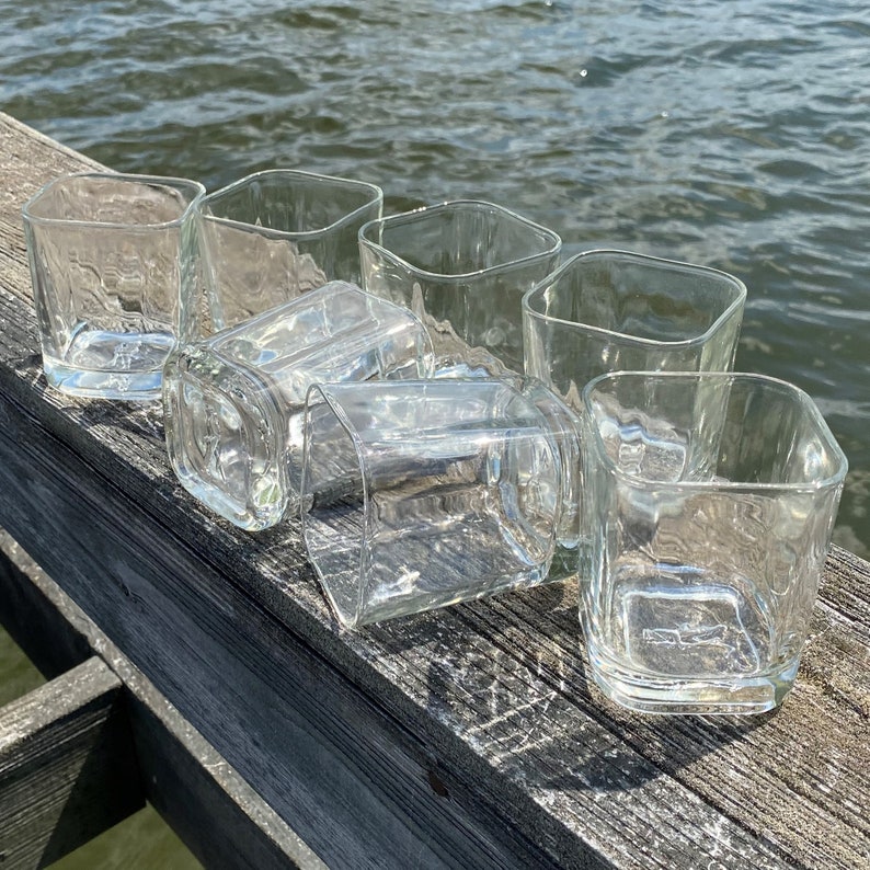 May include: Six clear glass tumblers with a textured surface, arranged in a row on a wooden surface.