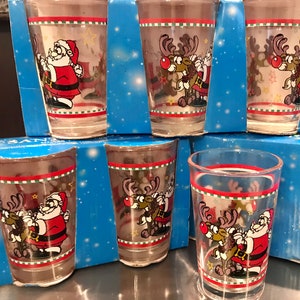 May include: Set of six clear shot glasses with Christmas-themed designs. Each glass features a red and green striped border and illustrations of Santa Claus and a reindeer. The glasses are displayed on blue boxes.