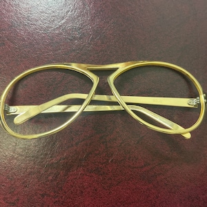 May include: Gold-tone aviator eyeglasses with a double bridge and light-colored temples. The glasses have a classic design, with a rectangular shape and a sleek, modern look. The frame is made of metal and has a shiny finish.
