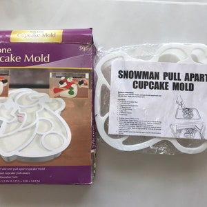 Puede incluir: Un molde para cupcakes de silicona blanco con forma de muñeco de nieve y su embalaje. La caja dice "Silicone Cupcake Mold" y "Snowman Pull Apart Cupcake Mold". El molde está diseñado para hacer 12 cupcakes individuales separables. Mide 27,9 cm x 22,8 cm x 3,8 cm.