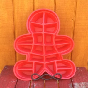 May include: A vibrant red gingerbread man-shaped baking mould with numerous square and rectangular compartments. The mould is made of a flexible material and rests on a decorative metal stand. The background is an orange wooden surface.