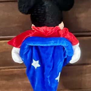 May include: A plush Mickey Mouse figure wearing a red cape and a blue wizard's hat with white stars. The figure has black ears and white gloves. The toy is designed to hang.