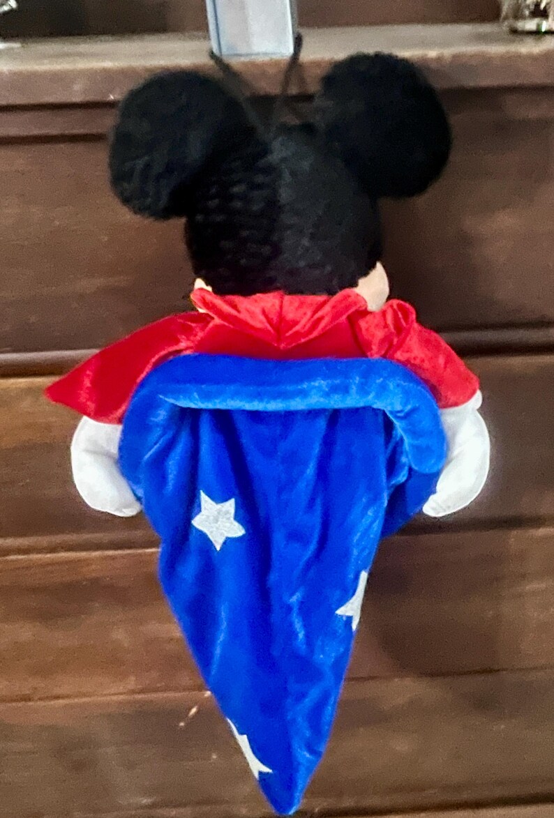 May include: A plush Mickey Mouse figure wearing a red cape and a blue wizard's hat with white stars. The figure has black ears and white gloves. The toy is designed to hang.
