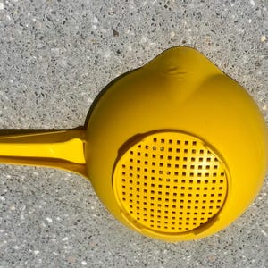 May include: A bright yellow plastic colander featuring a round bowl with a perforated base and a long, flat handle with a small circular hole at the end for hanging. The kitchen tool has a smooth, solid finish and a simple, functional design.