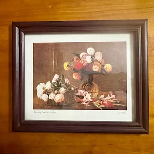 May include: A framed print depicting a floral still life. The image shows a vase of mixed flowers in shades of red, yellow, and white. A separate arrangement of white roses and pink gladioli is also visible. The frame is a dark brown colour.