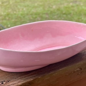 Op de afbeelding: Een lichtroze keramische schaal met een gebogen, bootachtige vorm. De schaal heeft een gladde, glanzende afwerking en staat op een kleine, afgeronde basis. De kom is leeg en lijkt vintage te zijn.