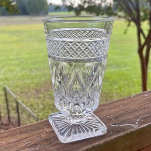 May include: A clear glass goblet featuring a textured diamond pattern around the upper rim and a vertical leaf-like motif on the main body. The vessel stands on a square base with a radiating sunburst design etched into the glass surface.