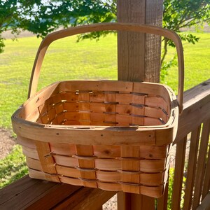 May include: A light brown, rectangular woven basket with a curved handle. The basket is made of thin strips of wood, woven together to create a sturdy container. The handle is attached to the sides of the basket.