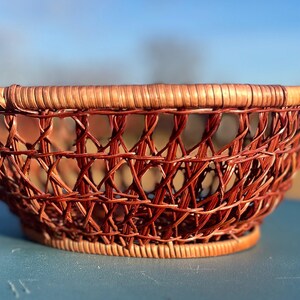 May include: A round, woven basket with a reddish-brown hue. The basket features an open, lattice-like design and a sturdy rim. The basket is likely made of natural materials and is suitable for storage or display.