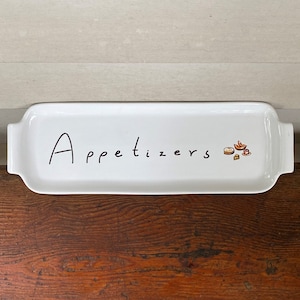 Puede incluir: Plato rectangular blanco con la palabra "Appetizers" en escritura negra. El plato tiene pequeñas asas en cada extremo y una pequeña ilustración de aperitivos en el lado derecho.