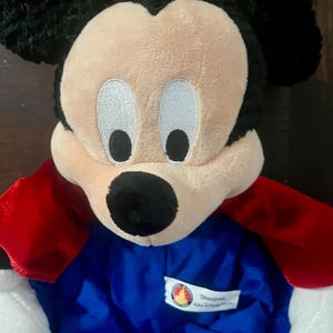 May include: A plush Mickey Mouse doll with a black head and ears, a tan face, and white eyes. The doll wears a blue outfit with red cape-like sleeves. A Disneyland tag is visible on the outfit.