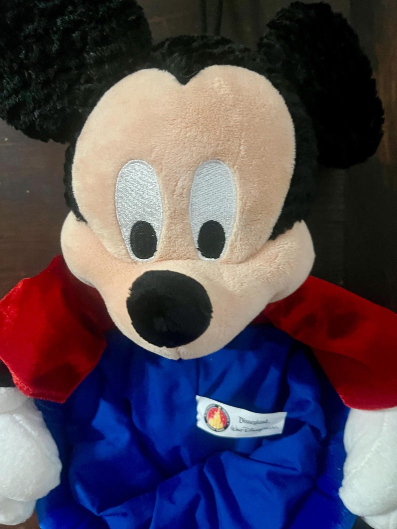 May include: A plush Mickey Mouse doll with a black head and ears, a tan face, and white eyes. The doll wears a blue outfit with red cape-like sleeves. A Disneyland tag is visible on the outfit.