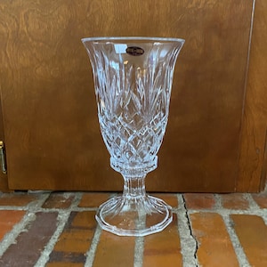 Op de afbeelding: Een heldere glazen vaas met een geslepen kristallen ontwerp. De vaas heeft een brede opening en een hoge, slanke voet. De vaas staat op een bakstenen oppervlak.