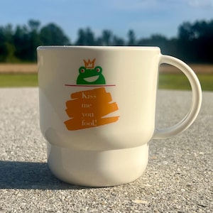 May include: A white ceramic mug featuring a graphic of a green frog wearing an orange crown. Below the frog, an orange brushstroke design contains the text "Kiss me you fool!" in white lettering. The mug has a smooth, rounded handle and base.