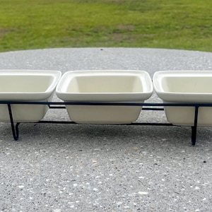 Pode incluir: Um conjunto de três taças quadradas de cerâmica em tom esbranquiçado dispostas em fila, mantidas dentro de um suporte de arame metálico preto. O suporte possui uma estrutura fina com pequenas alças nas extremidades e quatro pés curtos.