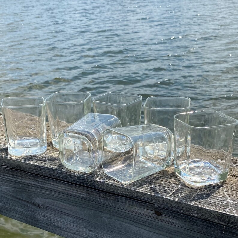 May include: Eight clear glass tumblers with a square shape, arranged on a wooden surface. The glasses are empty and appear to be made of thick glass.