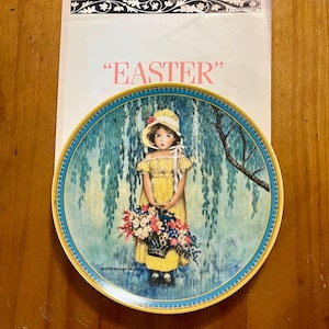 Puede incluir: Un plato decorativo con una niña con un vestido y sombrero amarillos sosteniendo una cesta de flores. El plato tiene un borde azul y amarillo y un fondo de sauces llorones. La palabra "EASTER" está impresa encima del plato.