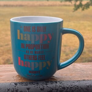 May include: A teal ceramic mug with a white interior and a curved handle. The mug features the text "ONE IS ONLY happy IN PROPORTION AS HE MAKES OTHERS FEEL happy" in various colors. The Hershey's logo is at the bottom.