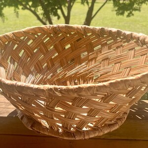 May include: A woven basket with a wide, shallow bowl shape. The basket is made of light brown woven material, with a textured surface. The basket is sitting on a wooden surface, with a green background.
