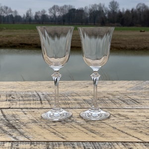May include: Two clear, stemmed wine glasses with a textured, fluted design on the bowl. The stems have a decorative, diamond-shaped element. The glasses are set on a weathered, wooden surface, with a blurred outdoor background.